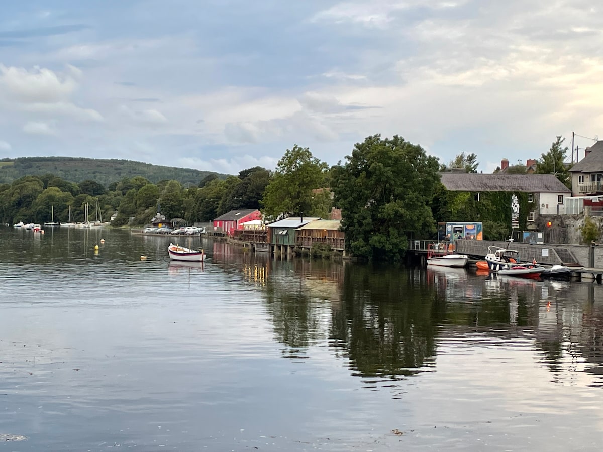 A calm river scene is depicted with moored boats along the water's edge. Lush greenery frames the river, while colorful buildings line the shore, reflecting in the still water. The sky, lightly clouded, hints at the serenity of the coastal environment.