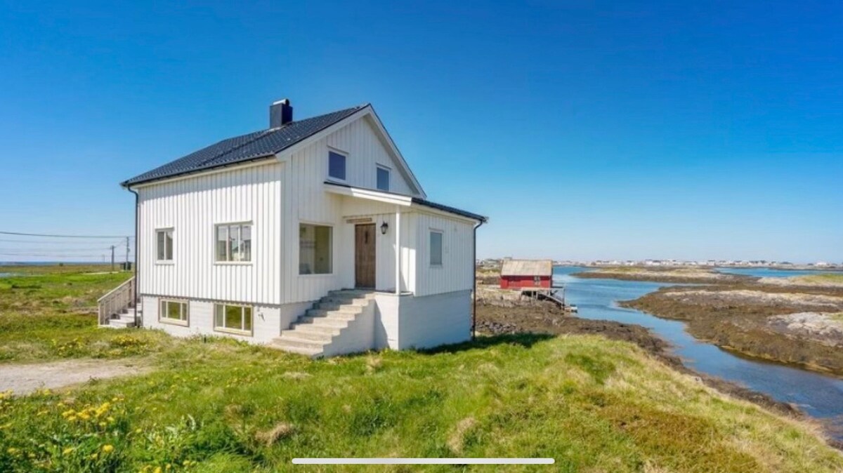 A two-story white house is positioned near a tranquil waterway, surrounded by greenery and wildflowers. The building features a black roof and large windows, allowing for natural light. Steps lead to the front entrance, while a rustic red structure is visible in the distance.