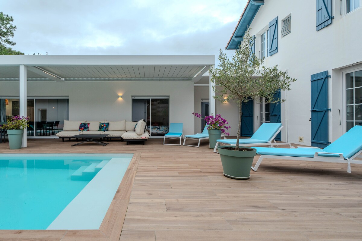 An outdoor terrace features a private heated pool surrounded by wooden decking. Lounge chairs with aqua blue cushions are positioned for relaxation. A shaded pergola offers a comfortable seating area with plush cushions, complemented by potted plants with vibrant flowers.