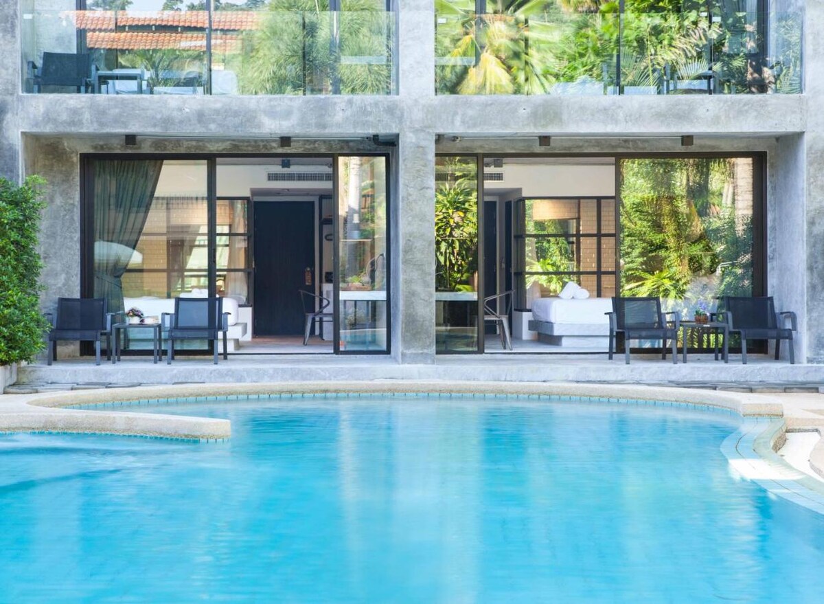 The image depicts a swimming pool with a smooth, rounded edge reflecting sunlight. Two spacious rooms are visible through large glass doors, showcasing comfortable seating and a well-lit interior with greenery in the background.