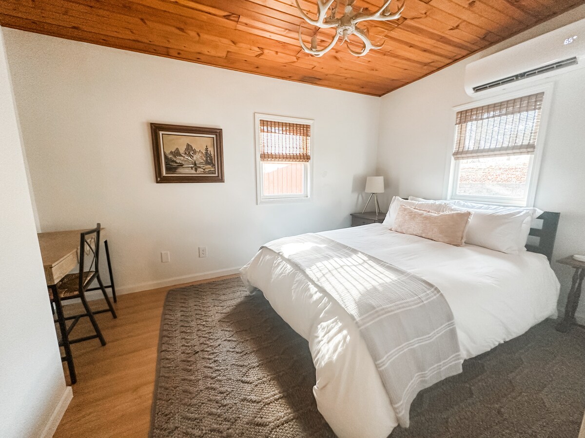 Ძირითადი საძინებელი 152 × 203 სმ ზომის საწოლით. Ანტლერის სტილის ჭაღი, სამუშაო სივრცე და ორიგინალური ხელოვნების ნიმუშები სრულდება ოთახს. 