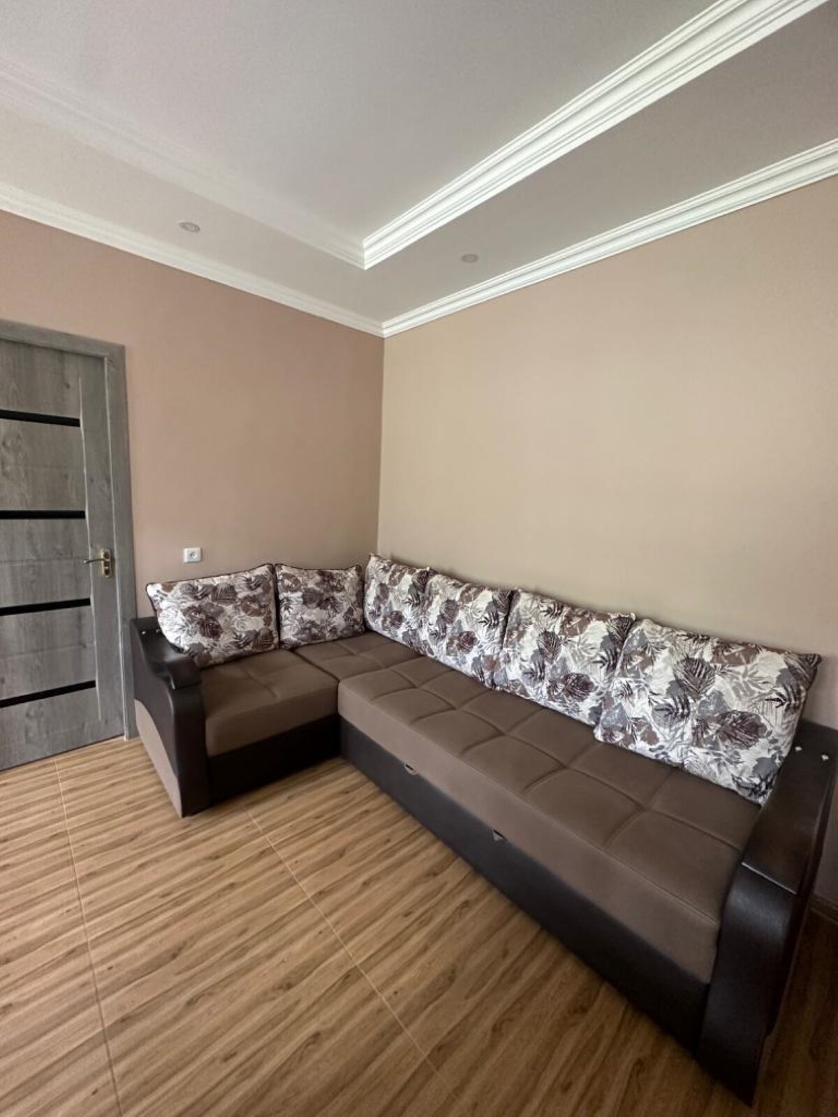 A comfortable seating area features a spacious sectional sofa with floral-patterned cushions. The neutral walls are complemented by a simple ceiling design, while natural light enhances the inviting space. A door is visible in the background, adding functionality to the room.