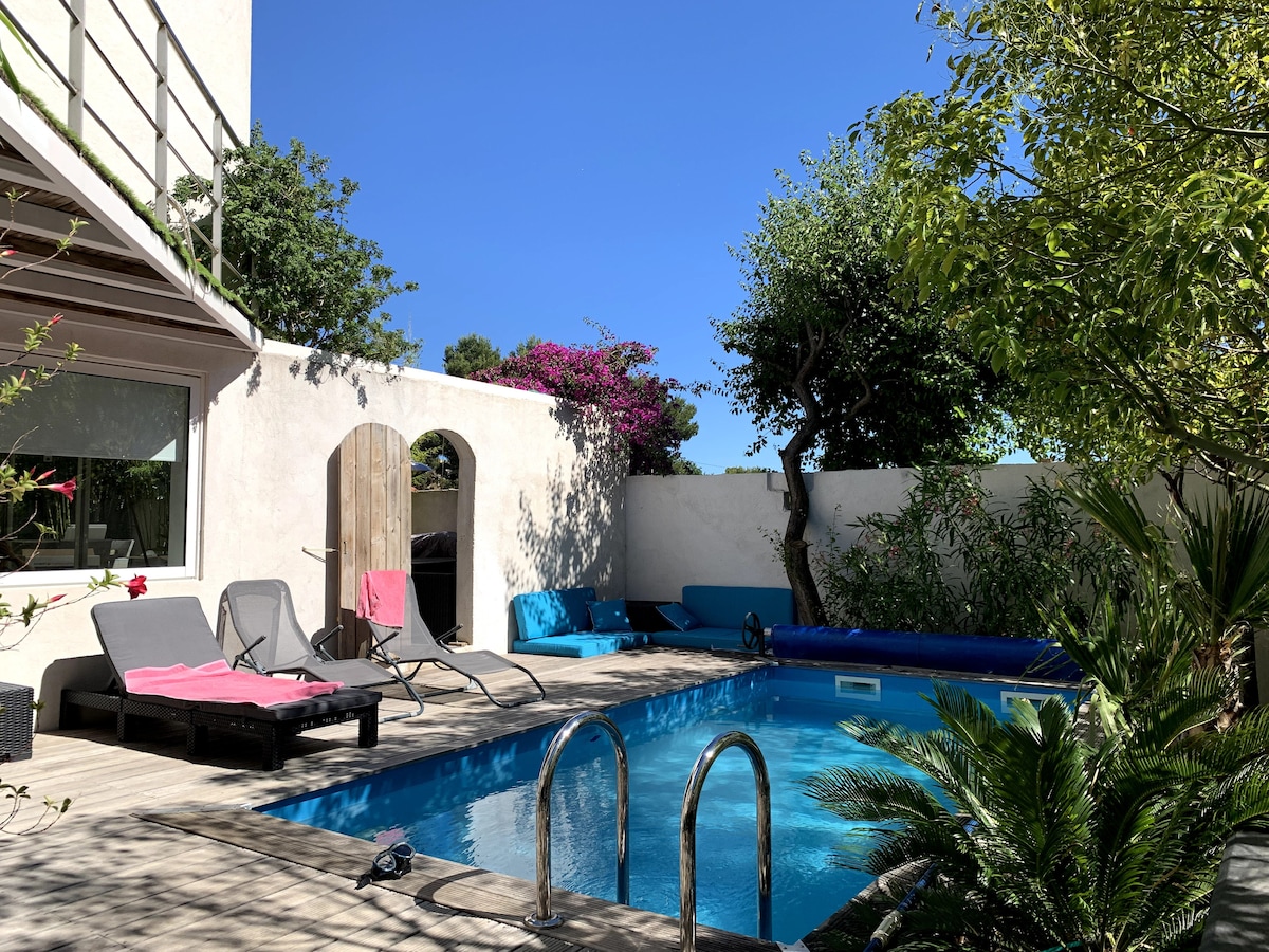 A private swimming pool is framed by green foliage and bright flowers. Sun loungers are positioned nearby, inviting relaxation. A sunny wooden deck surrounds the pool, with vibrant colored cushions placed against the wall, enhancing the outdoor space's tranquil ambiance.