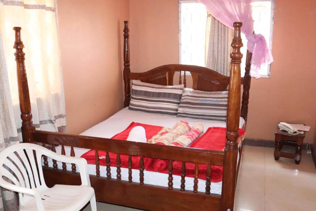 A spacious bed with a wooden frame is positioned against a warm-toned wall. Soft linens and towels are neatly arranged on the bed. A plastic chair and a small side table are visible, along with curtains that gently frame the windows.
