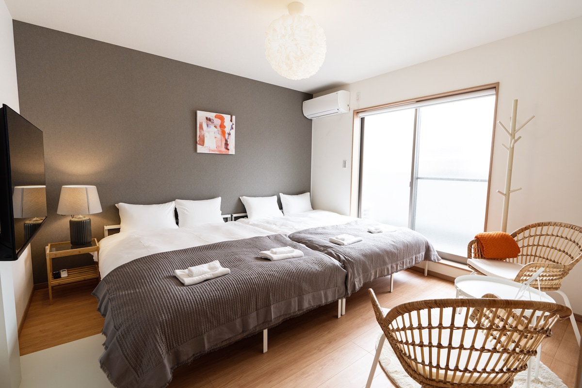 A spacious sleeping area is furnished with two large beds, each topped with white linens and large towels. A modern chair and small table complement the room, which features a large window that allows natural light to illuminate the space.