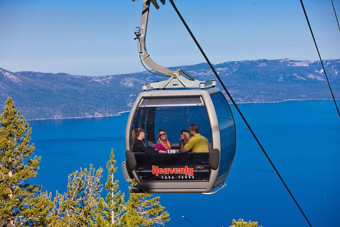 A gondola cabin is elevated above the landscape, carrying passengers amidst stunning views of blue waters and mountains in the background. The clear sky enhances visibility of the scenic surroundings, creating an ideal opportunity for sightseeing during the ride.