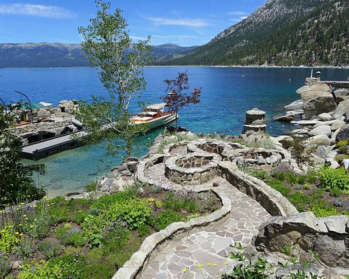 A landscaped pathway winds through vibrant flowers, leading toward the clear blue waters of Lake Tahoe. Natural rock formations are positioned along the shoreline, and a boat can be seen moored at the dock, surrounded by scenic mountain views in the background.