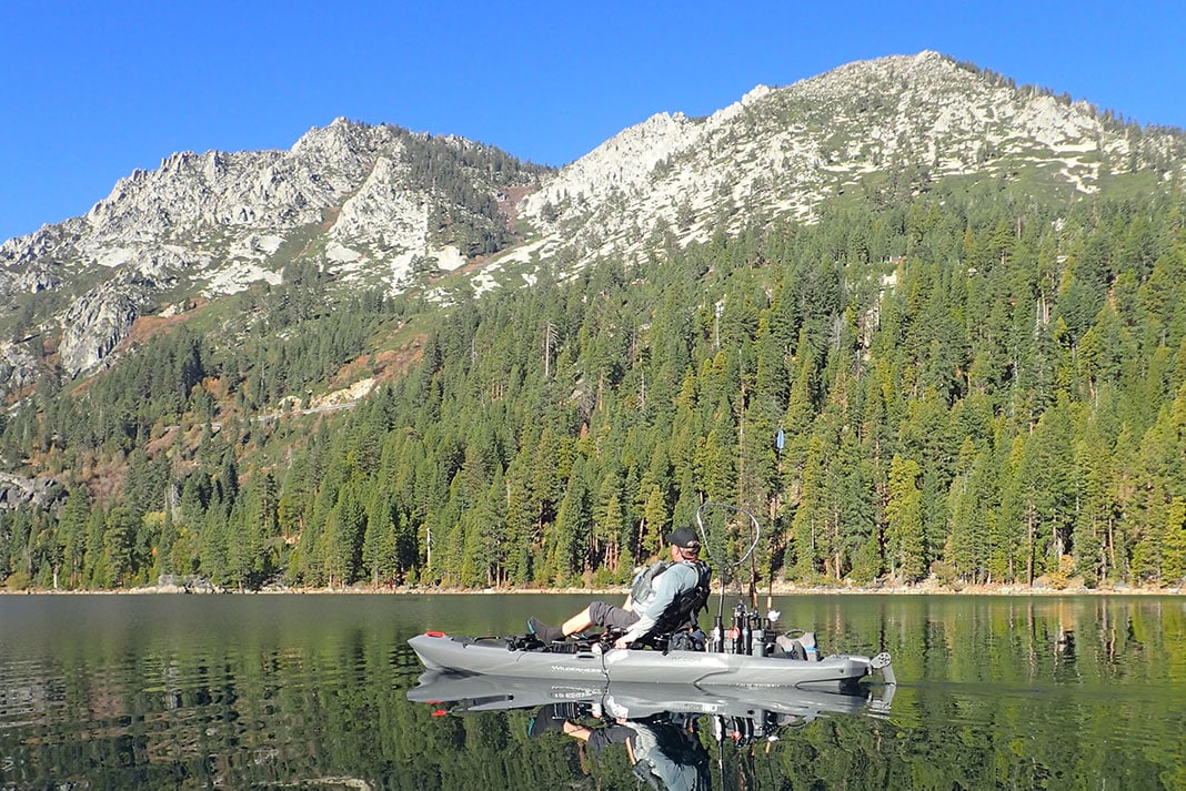 A person is seen fishing while seated in a kayak, surrounded by calm waters reflecting the surrounding mountains and dense pine trees. The clear blue sky adds to the serene atmosphere of the lake.
