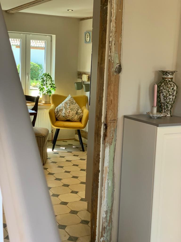 A glimpse into the cozy cottage reveals a light-filled dining area featuring a yellow accent chair with a floral cushion. The hexagonal tiled floor creates a stylish backdrop, while a vase and candle add subtle decorative touches nearby.