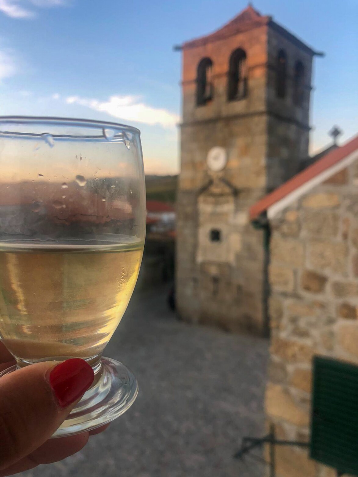 A clear glass holds a light-colored drink in focus, with a stone building featuring a clock tower in the background. The rustic architecture and gravel pathway are softly illuminated by the evening light, creating a serene outdoor setting.