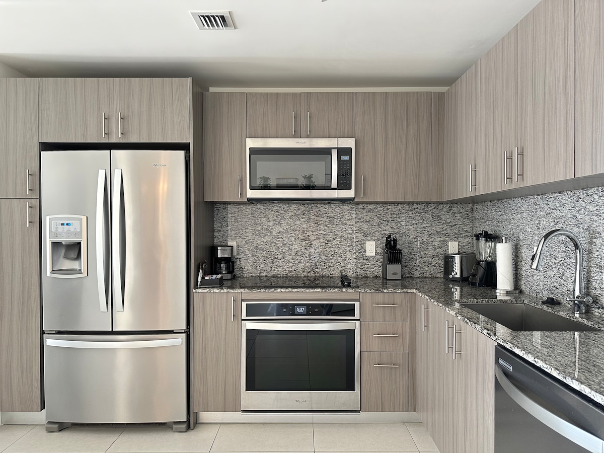 The kitchen showcases modern stainless steel appliances, including a refrigerator, microwave, and oven. Elegant cabinetry complements the granite countertops, and a coffee machine and toaster are positioned for convenience. A sleek sink is installed beside a textured backsplash, adding a stylish touch to the functional space.