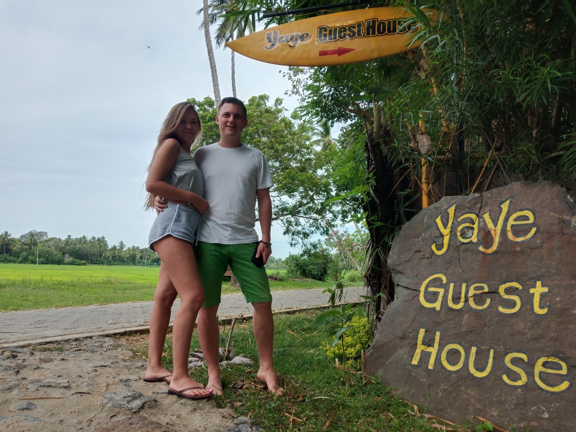 A sign displaying 'Yaye Guest House' is prominently positioned on a large rock, complemented by a vibrant surfboard above. Lush greenery and palm trees frame the scene, while a nature path leads into the distance, creating a welcoming entrance to the guest house.