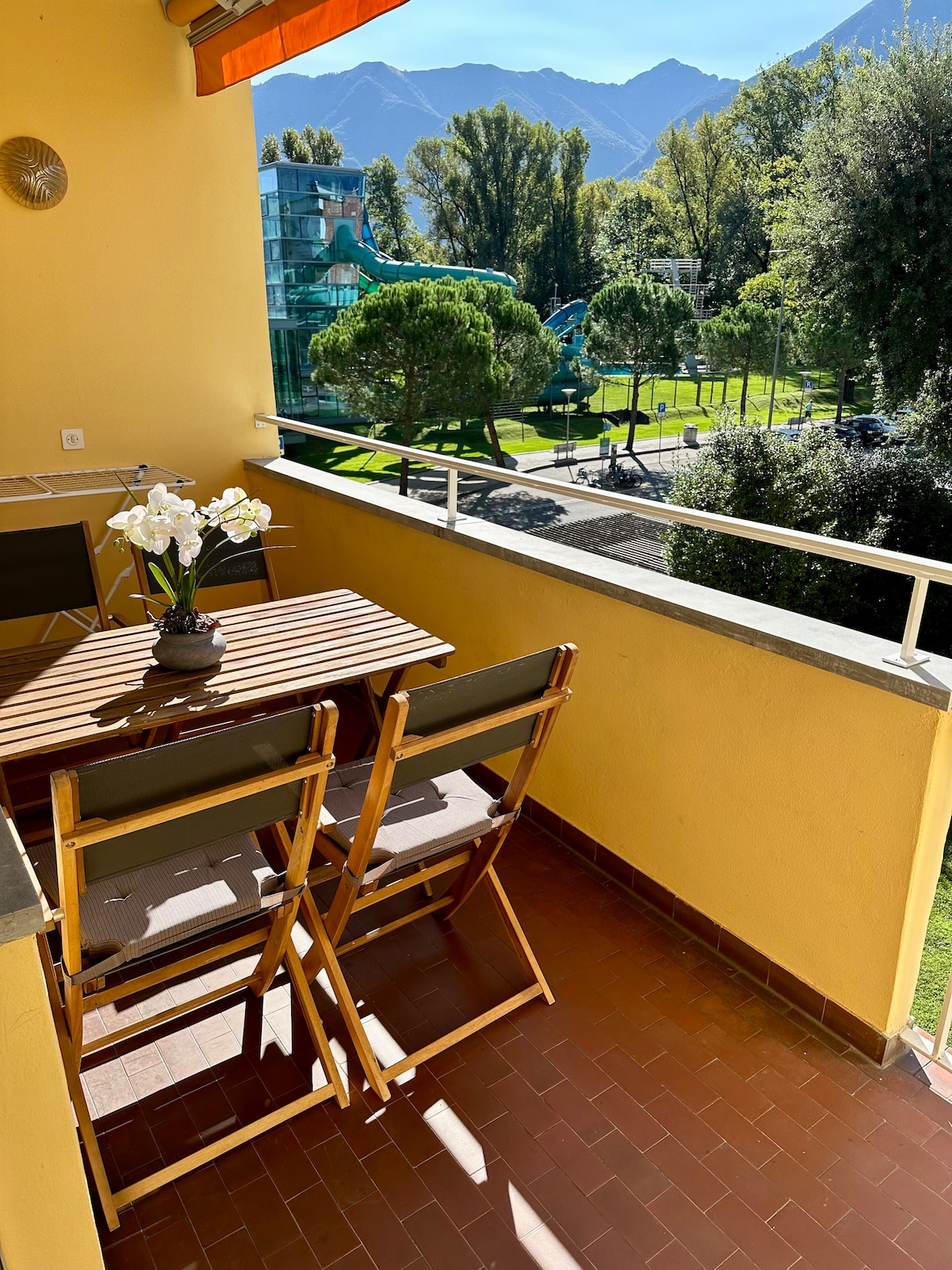 A sunny balcony area features a wooden table and two folding chairs, offering a relaxing space for outdoor dining. The bright yellow wall complements the surrounding greenery and distant mountains visible in the background, creating a tranquil environment.