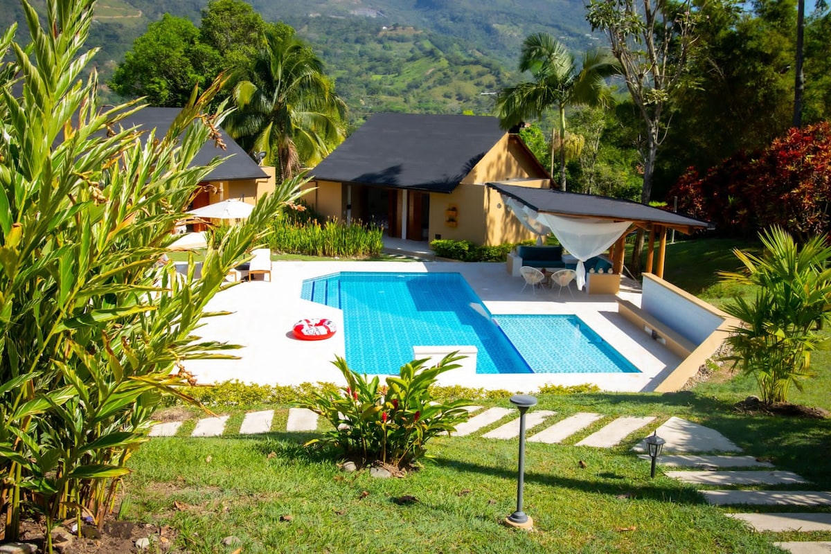 A spacious outdoor area features a clean, inviting pool bordered by lush greenery. Sun loungers are positioned next to the water, and a shaded seating area provides a comfortable space for relaxation. The backdrop showcases gently rolling hills under a clear sky.