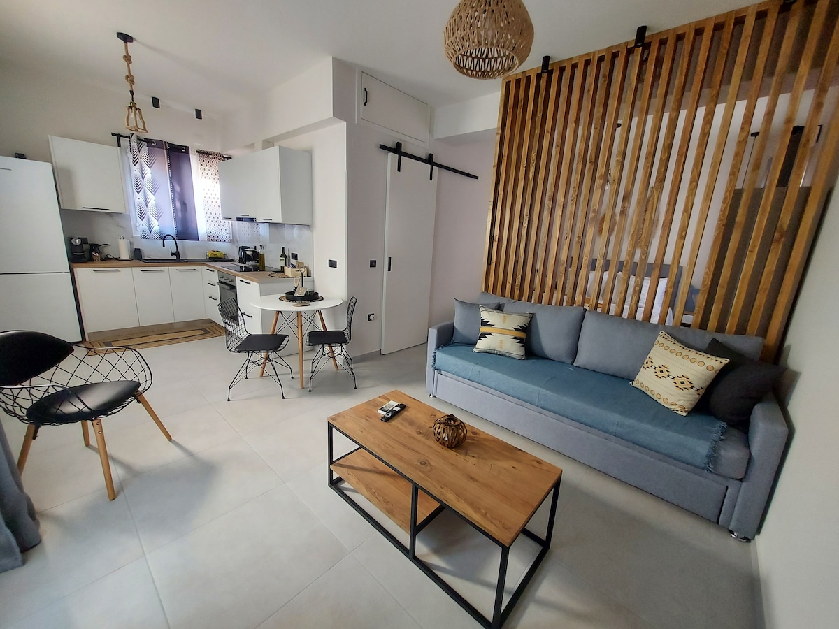 A modern living area is presented with a gray couch and a wooden coffee table. A black dining set is set for two, complementing the kitchen in the background. Natural light streams through a window, enhancing the ambiance of the open space.