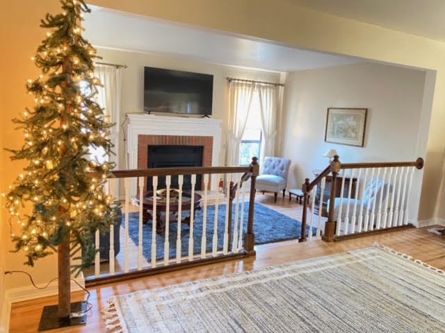 A welcoming living area features a decorative illuminated tree beside a white railing, leading to a cozy seating arrangement with two armchairs. A classic coffee table is placed on a textured rug, with natural light filtering through the nearby window, highlighting the warmth of the space.