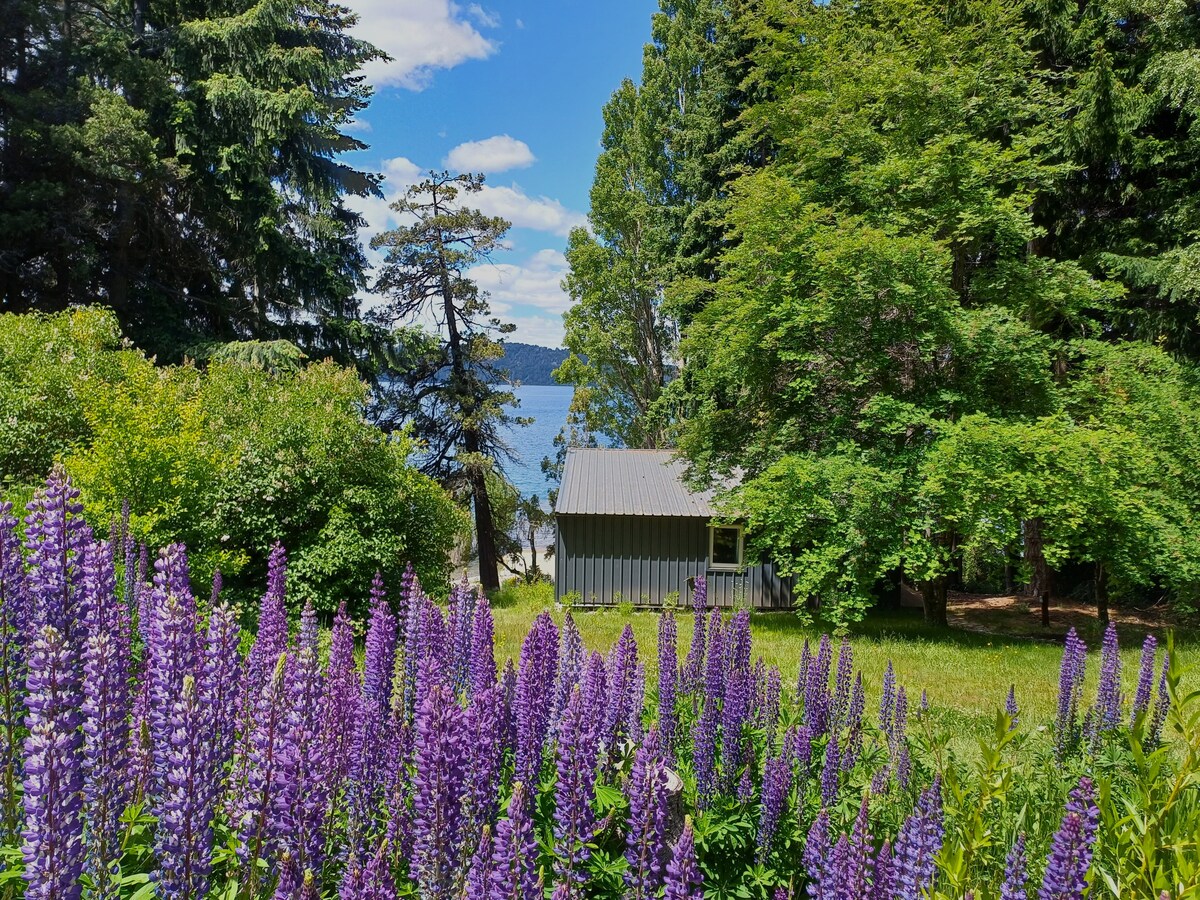 A serene view of a cabin surrounded by lush greenery, with vibrant purple flowers in the foreground. A glimpse of the lake is visible through the trees, framed by a clear blue sky dotted with clouds.