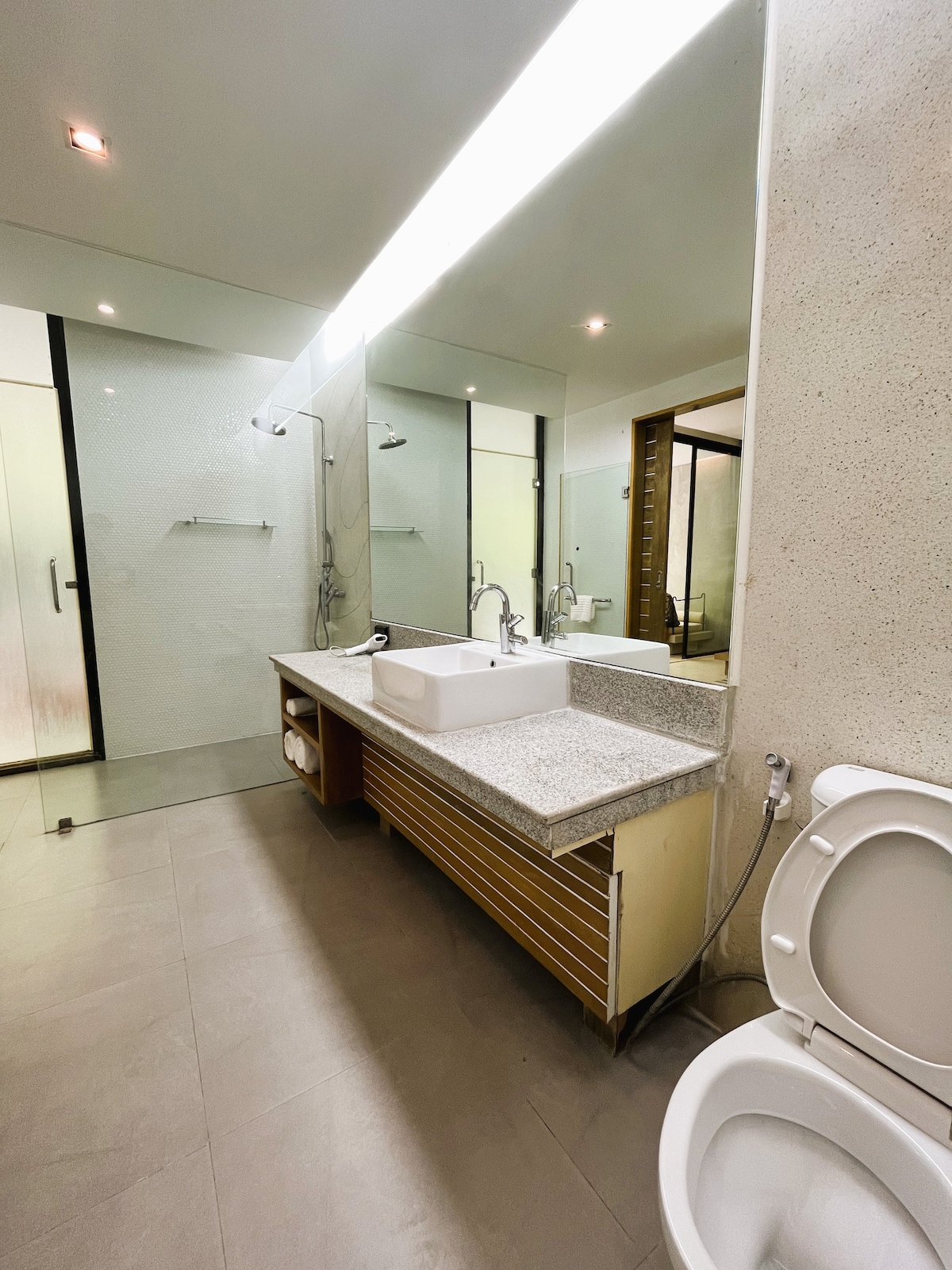 The bathroom is designed with a modern aesthetic, featuring a double sink on a granite countertop. A large mirror reflects the space, and the area is well-lit with overhead lighting. The shower area is enclosed, while additional fixtures are neatly arranged.