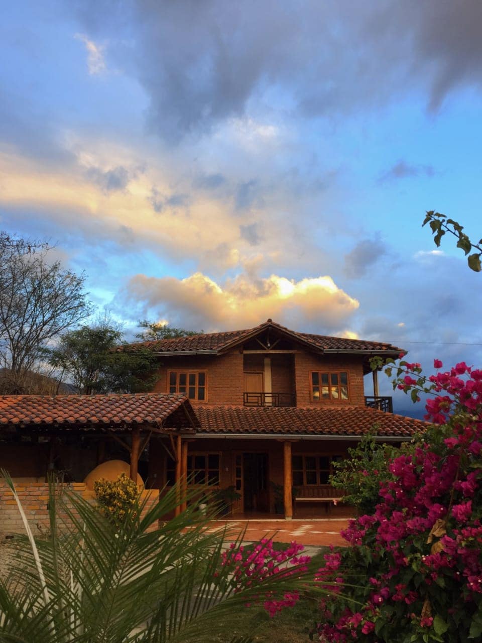 A two-story brick house is set against a sky filled with clouds. A covered porch extends from the lower level, and vibrant pink flowers and greenery are visible in the foreground. The architecture features a traditional design with a tiled roof and wooden accents.
