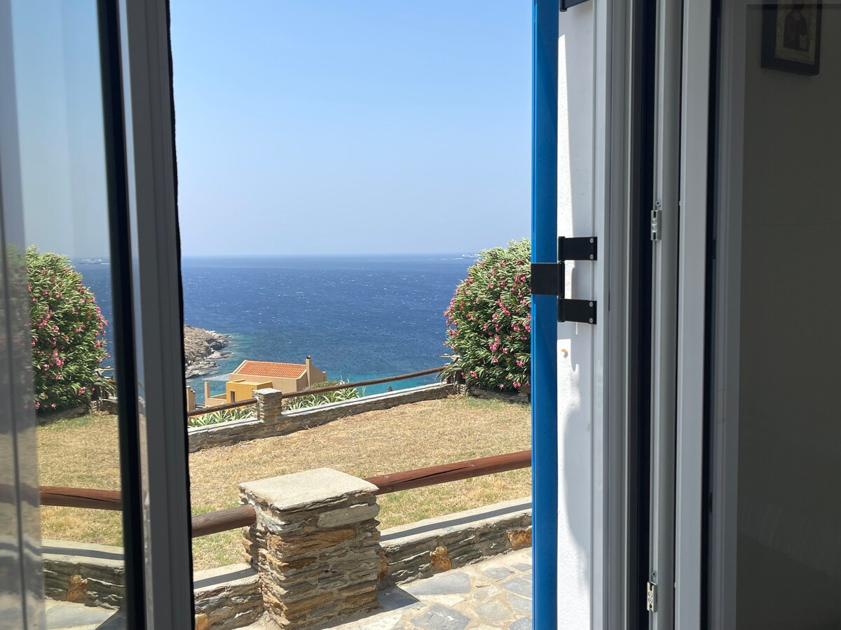 A view from an open door reveals a tranquil sea beyond a well-maintained garden. Lush greenery frames the foreground, contrasting with the deep blue water of the Aegean Sea. The clear sky enhances the inviting atmosphere of this coastal setting.