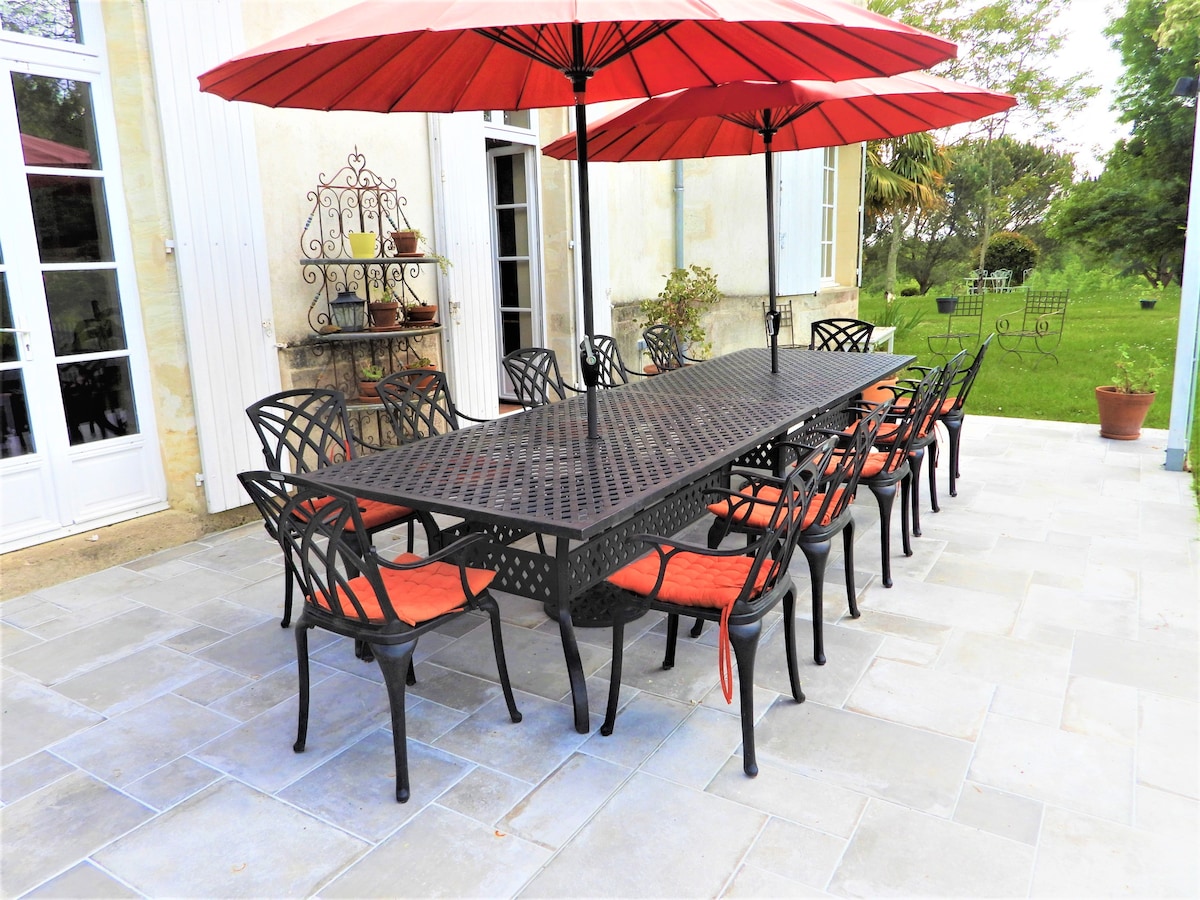 A large outdoor dining area is featured, with a long table surrounded by numerous black chairs with padded red cushions. Red umbrellas provide shade over the table. Natural light fills the space, with greenery visible in the background.