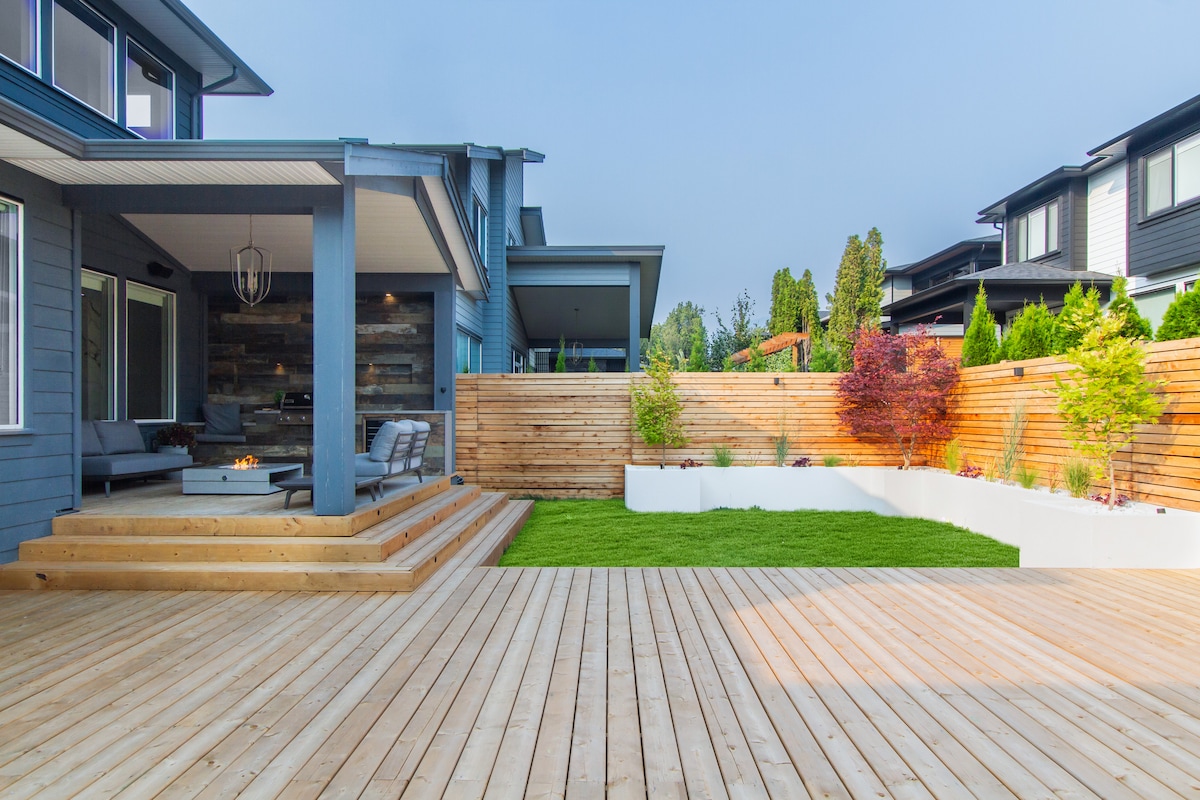 A spacious outdoor deck features a wooden surface with steps leading to a covered lounge area. Surrounding vegetation is highlighted by a mix of colorful plants and trees, while a low wall defines a raised garden area. Soft grass complements the deck, creating an inviting atmosphere.