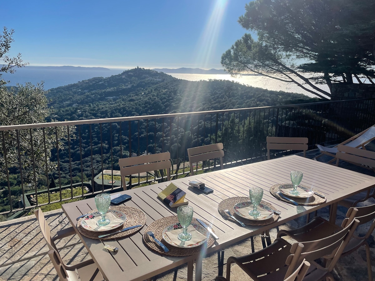 An outdoor dining area is set with place settings on a long table, surrounded by chairs. The landscape features rolling hills and a panoramic view of the sea, enhanced by natural light. Trees are visible in the foreground, framing the serene environment.
