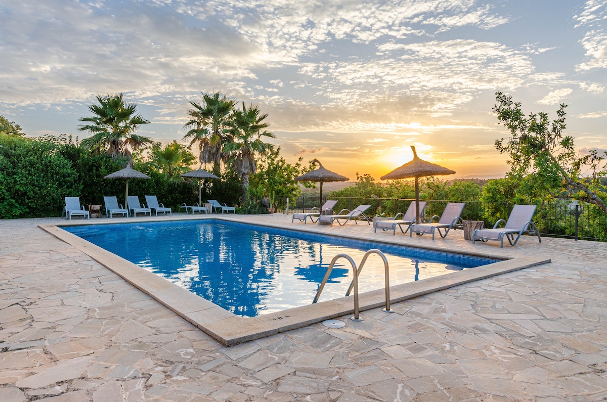 An outdoor swimming pool is surrounded by lounge chairs and sun umbrellas, with greenery in the background. The setting captures a sunset sky, reflecting warm hues across the water's surface. A stone patio frames the pool area, creating a spacious environment for relaxation.