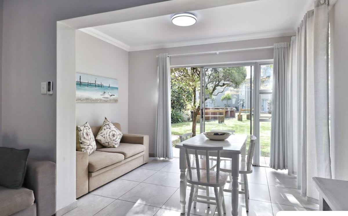 A bright dining area features a simple white table surrounded by four chairs. Natural light floods the space, illuminating the soft tones of the walls. A comfortable sofa is positioned nearby, and large windows offer views of the garden outside.