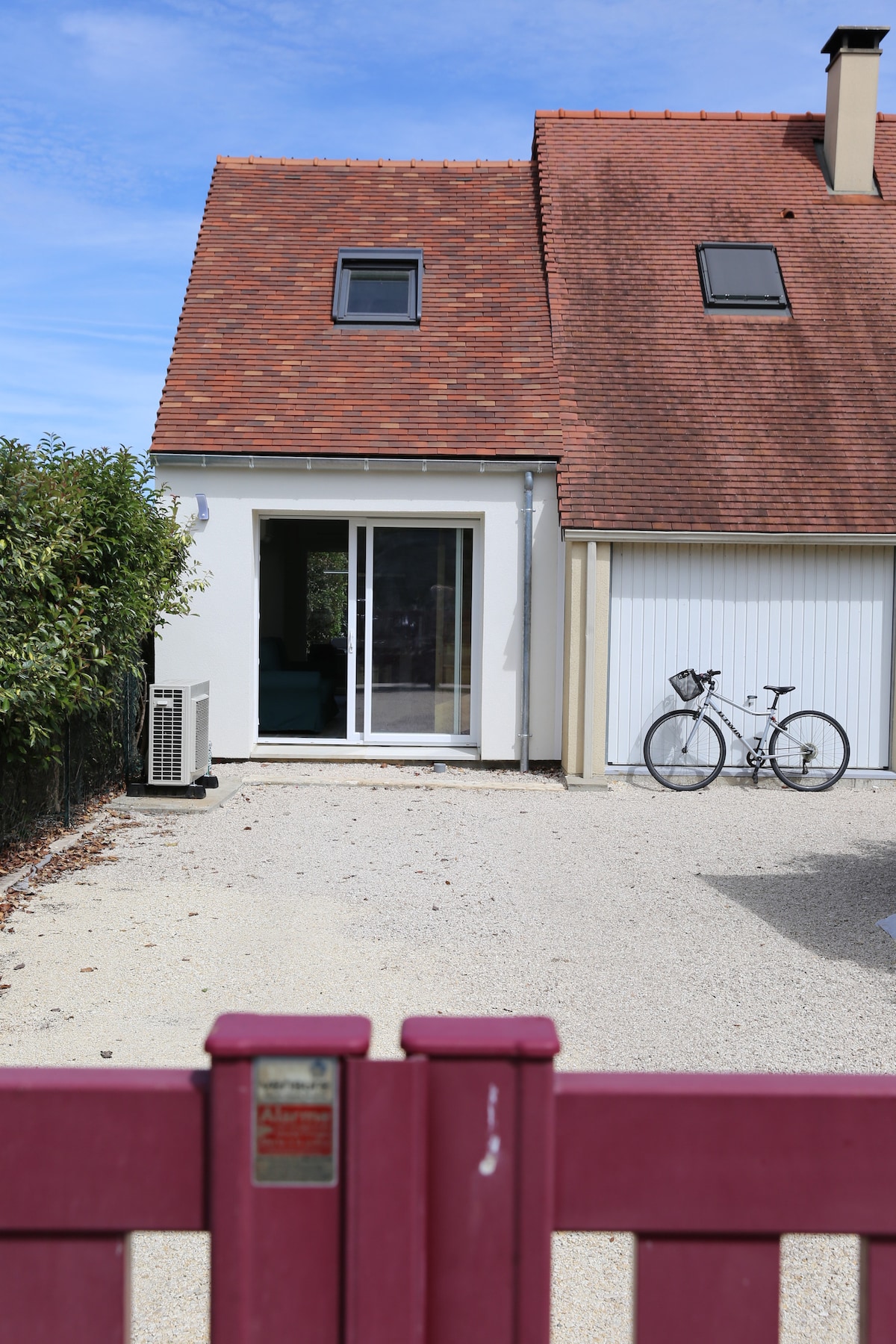 A view of the property entrance is presented, featuring a maroon gate leading to a gravel path. The exterior showcases a two-story building with a sloped roof and two small roof windows. A bicycle is parked next to the sliding glass door, which opens to the living area.