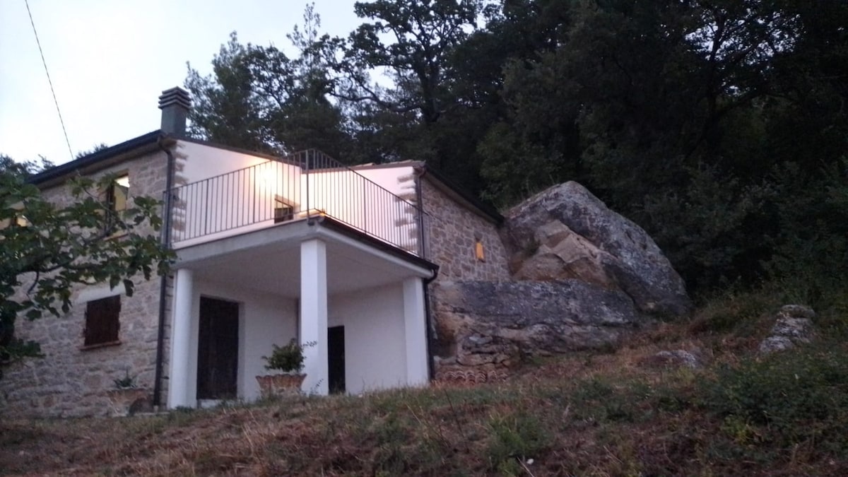 A rustic stone house is situated against a rocky landscape, highlighting a large boulder integrated into the structure. The property features a covered balcony with metal railings, while trees and greenery surround the area, enhancing the serene atmosphere of the countryside.