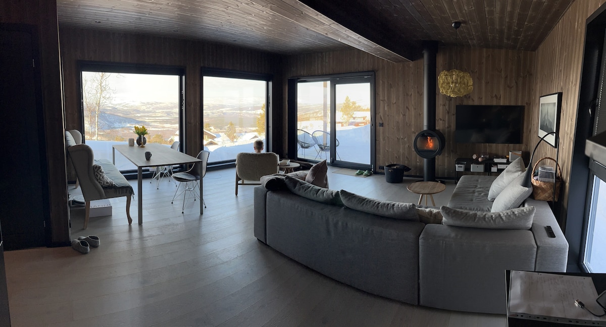 A spacious living area features large floor-to-ceiling windows that provide a panoramic view of the surrounding landscape. A cozy seating arrangement includes a sectional sofa and armchairs. A dining table is positioned near the window, complemented by a television and a wood stove.