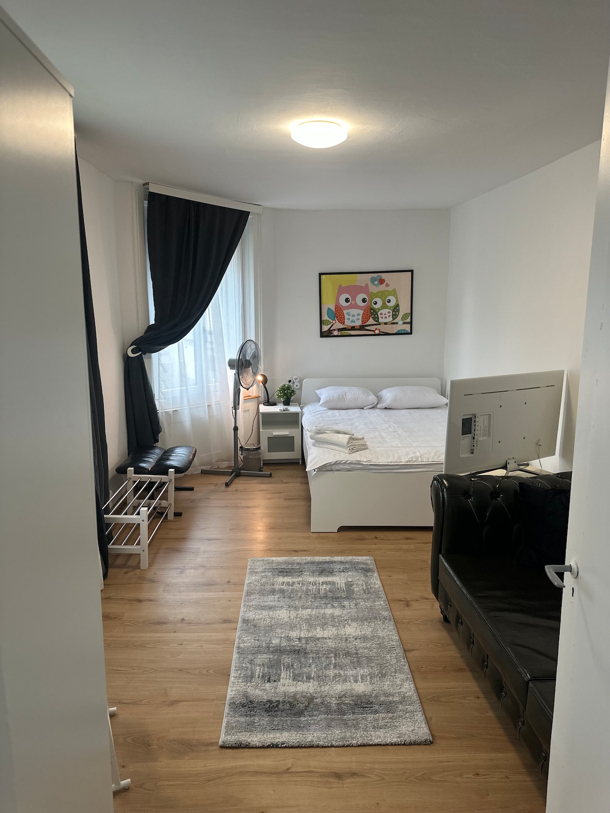 A neatly arranged bedroom features a spacious bed with crisp white linens. A colorful artwork hangs on the wall above, complemented by a floor fan and a small bedside table. Natural light filters through a window, with a dark curtain partially drawn.