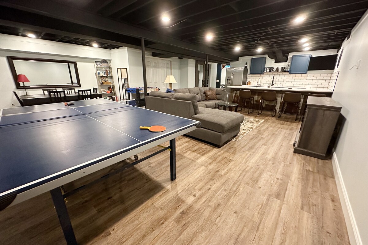 A finished basement is displayed, featuring a ping pong table on the left and a cozy seating area with a sectional sofa. In the background, a bar with high stools is visible, along with a well-lit space that promotes relaxation and entertainment.