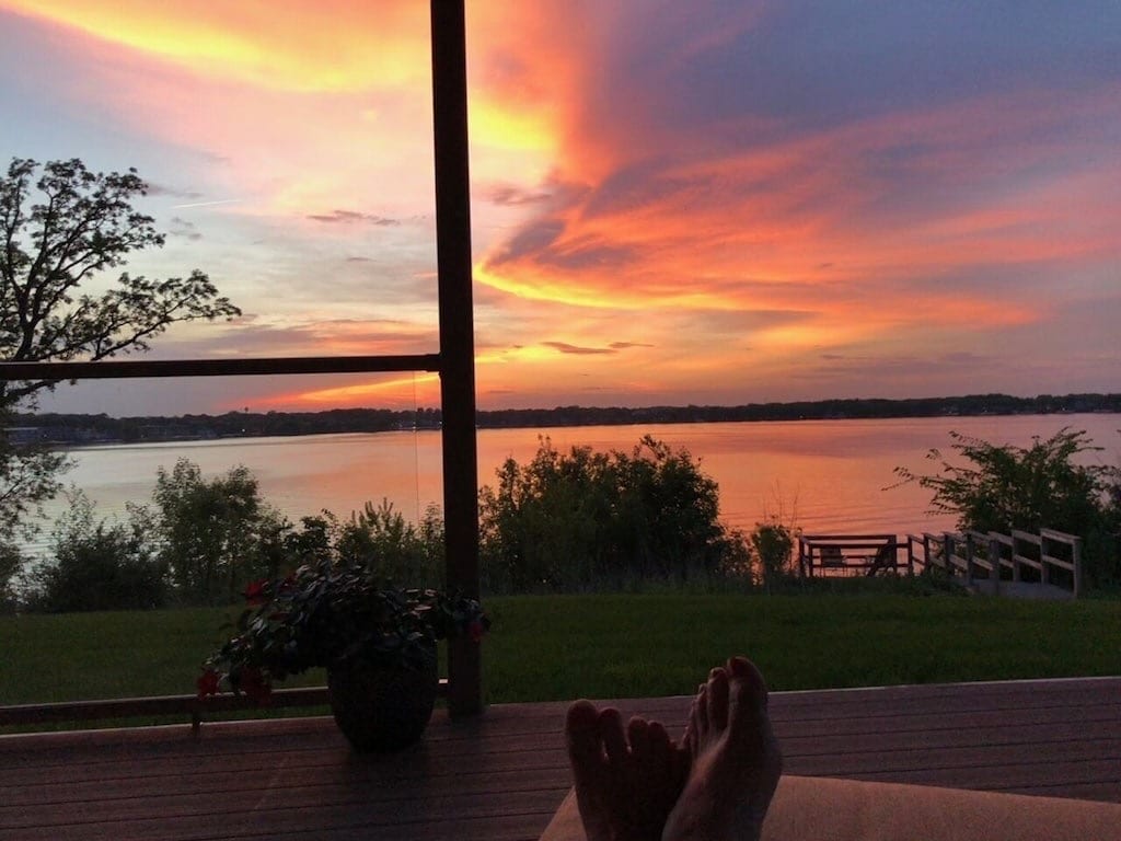 A serene view of a lake at sunset is captured from a private deck. Vibrant colors blend across the sky, reflecting on the water. Lush greenery and a small seating area are visible in the foreground, enhancing the natural setting.