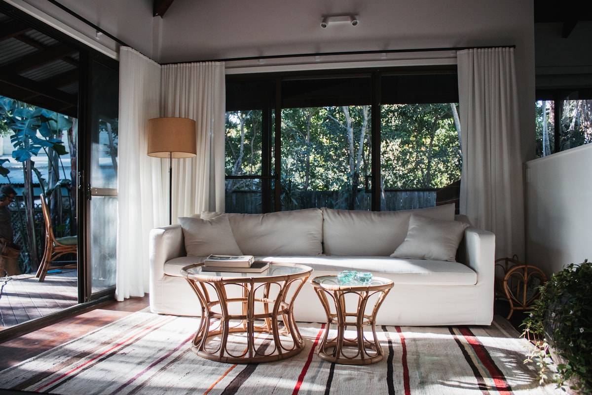A light-filled living area features a spacious white couch and two woven coffee tables, complemented by a cozy rug with a colorful pattern. Large glass doors provide outdoor views, while sheer curtains frame the windows, allowing natural light to enhance the atmosphere.