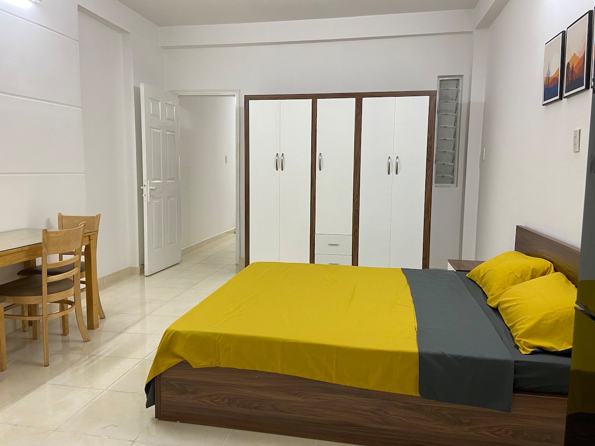 A spacious bedroom features a large bed with a yellow and gray bedding set. A wooden table and chairs are positioned nearby, along with a wardrobe with white doors. Soft lighting enhances the neutral-colored walls and flooring.