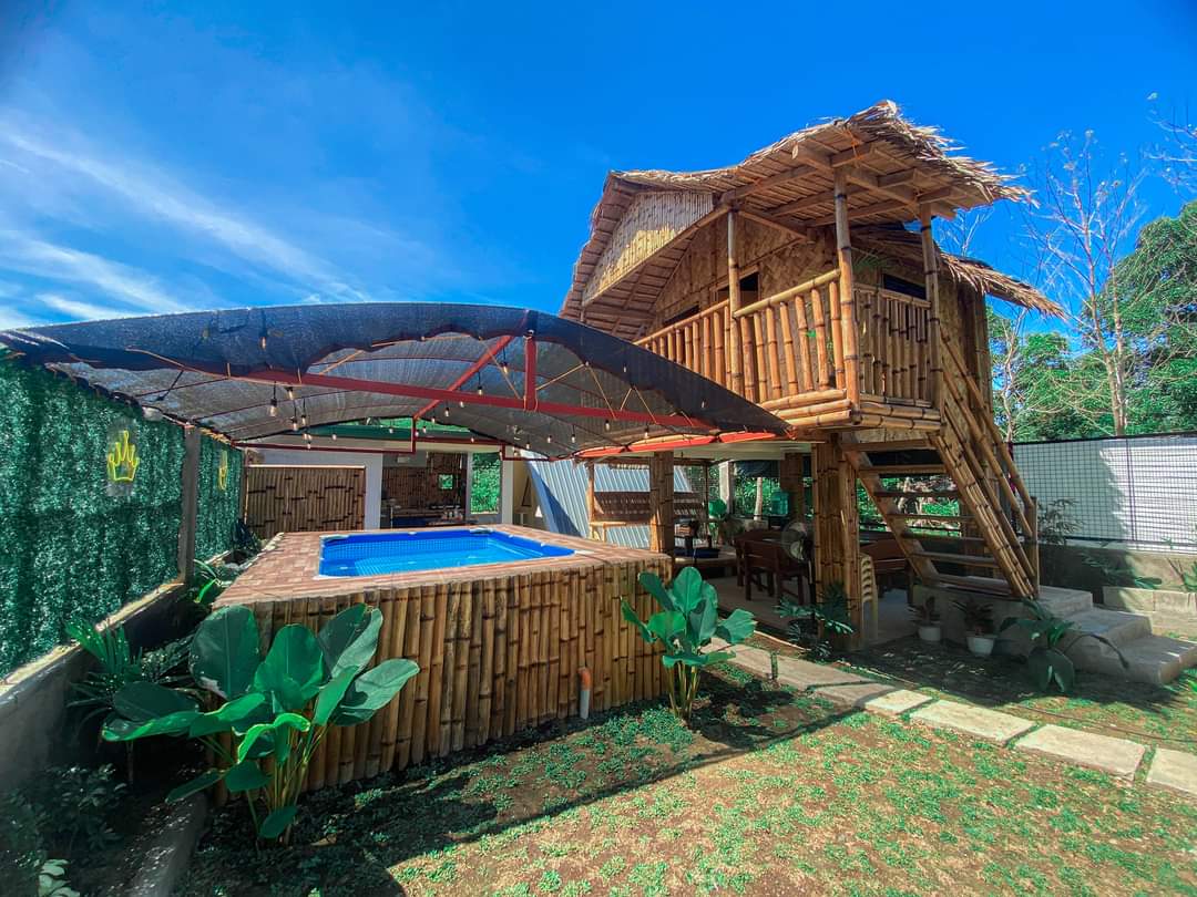 A bamboo structure features a covered deck that overlooks a small portable pool. Surrounding greenery and tropical plants border the area, while the clear blue sky provides a vibrant backdrop. The layout enhances the outdoor living experience, perfect for relaxation and gatherings.