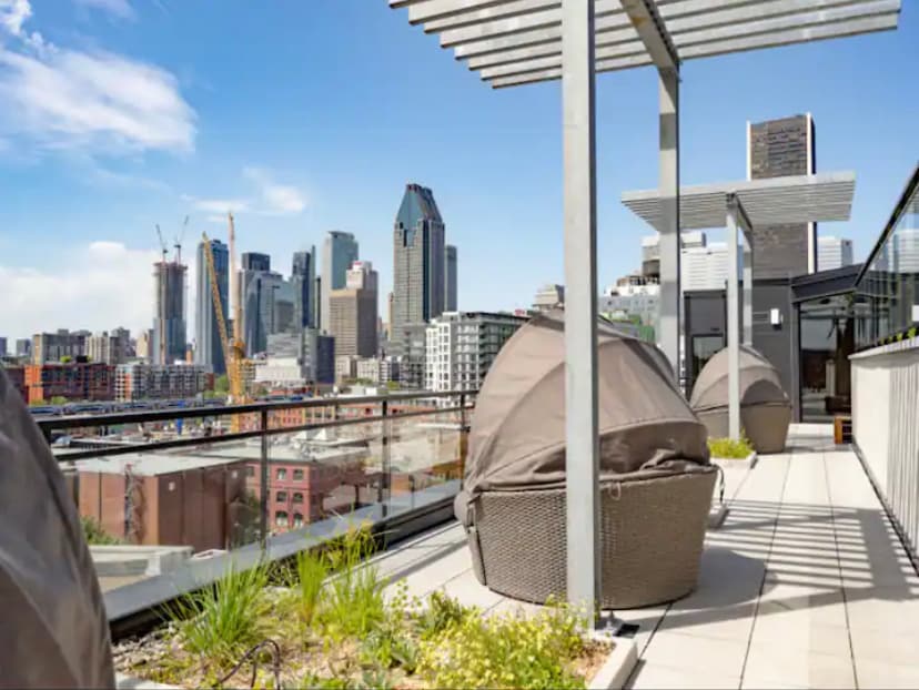 A spacious rooftop terrace is shown, featuring modern lounge pods positioned along the edges. The city skyline is visible in the background, with a clear sky contributing to the vibrant scene. Greenery interspersed throughout the space adds a touch of nature.