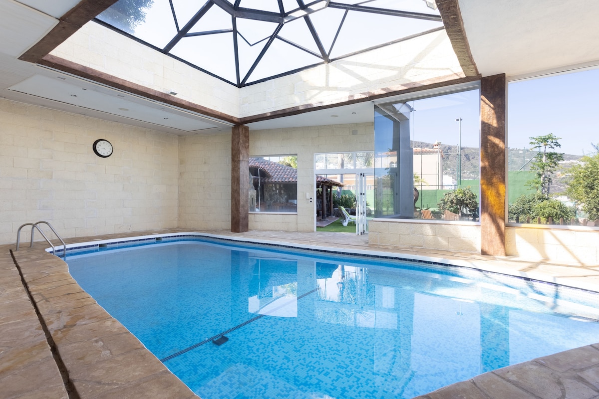 An indoor pool features clear blue water, surrounded by light stone walls and large windows that let in natural light. A transparent roof allows for a view of the sky, enhancing the atmosphere. A clock is mounted on the wall, indicating the time.