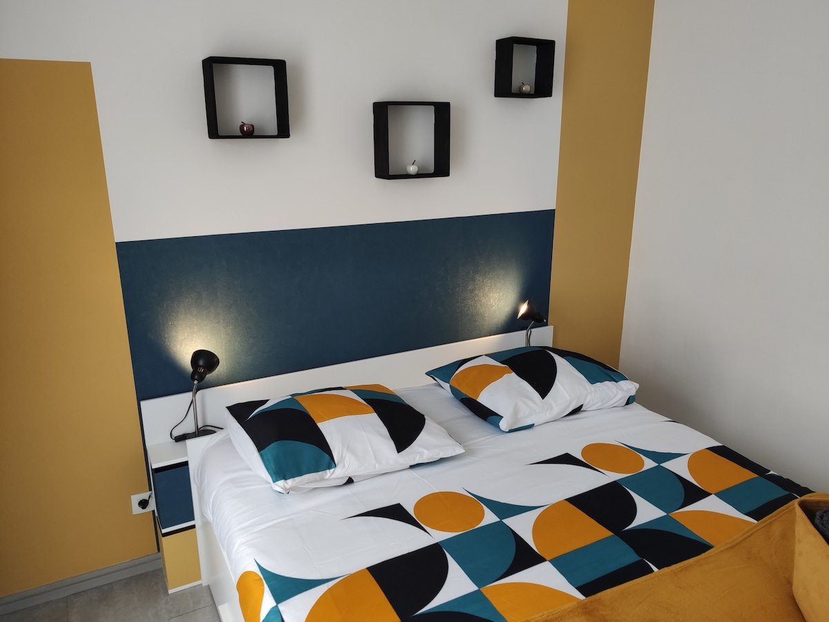 The sleeping area is presented with a vibrant geometric-patterned bedspread covering a spacious bed. Two bedside lamps provide focused lighting, while geometric shelves adorn the wall above, displaying small decorative items.