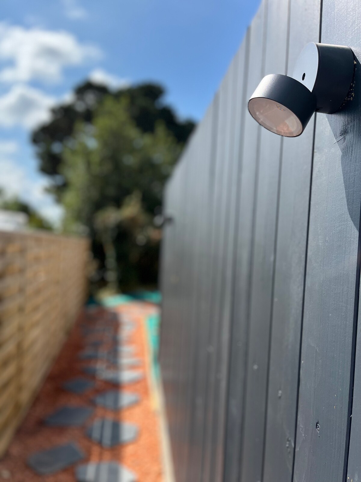 A side pathway is lined with stepping stones and pebbles, leading through a well-maintained outdoor space. A wooden fence with light fixtures is visible, alongside greenery from the surrounding garden, creating a private atmosphere for guests.