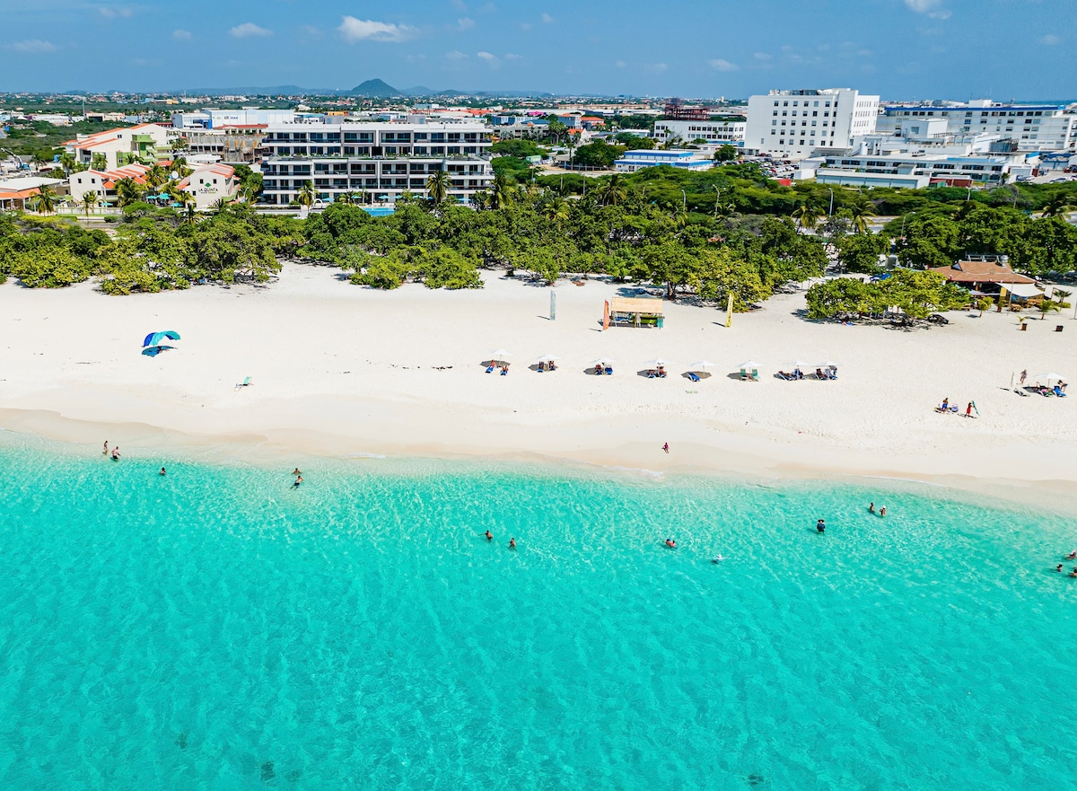 The image captures a scenic view of a sandy beach lined with vibrant turquoise waters. Beachgoers can be seen enjoying the shoreline, while umbrellas and beach chairs are strategically placed along the sand. The backdrop features several buildings and lush greenery, enhancing the tranquil atmosphere.