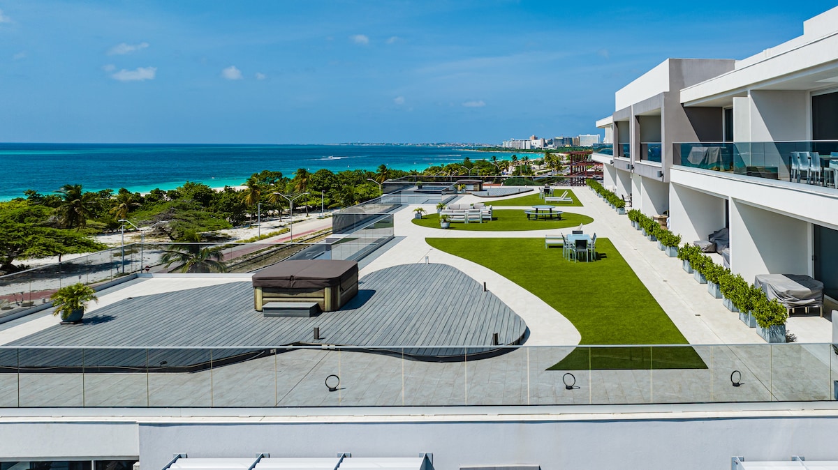 An expansive outdoor space showcases a tranquil layout with manicured grass and wooden decking. A Jacuzzi sits centrally, surrounded by lounge chairs and dining areas. The coastline can be seen in the background, featuring a clear blue sky and azure waters that reflect the sunlight.
