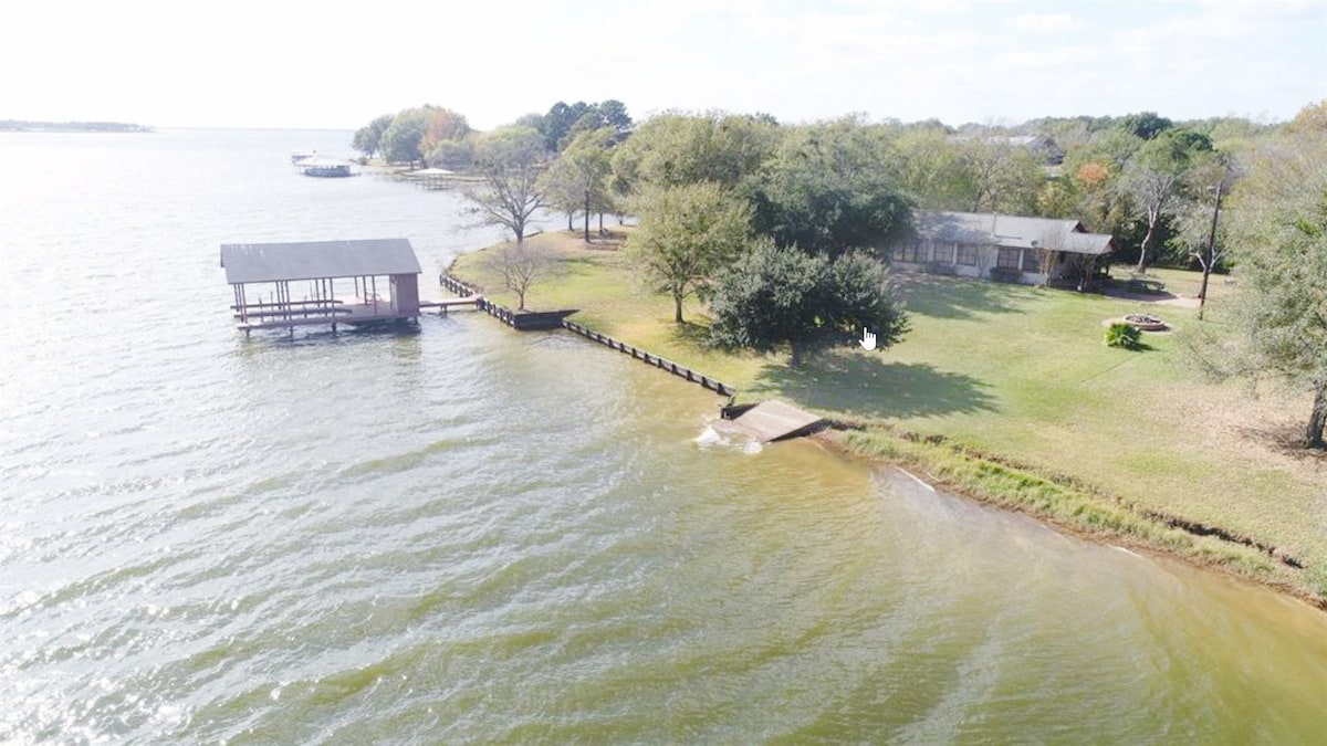 The image captures a serene waterfront view, showcasing a grassy area leading to the water. A covered dock is visible to the left, with trees dotting the landscape. The peaceful water reflects the sky, creating a calm ambiance enhanced by the natural surroundings.
