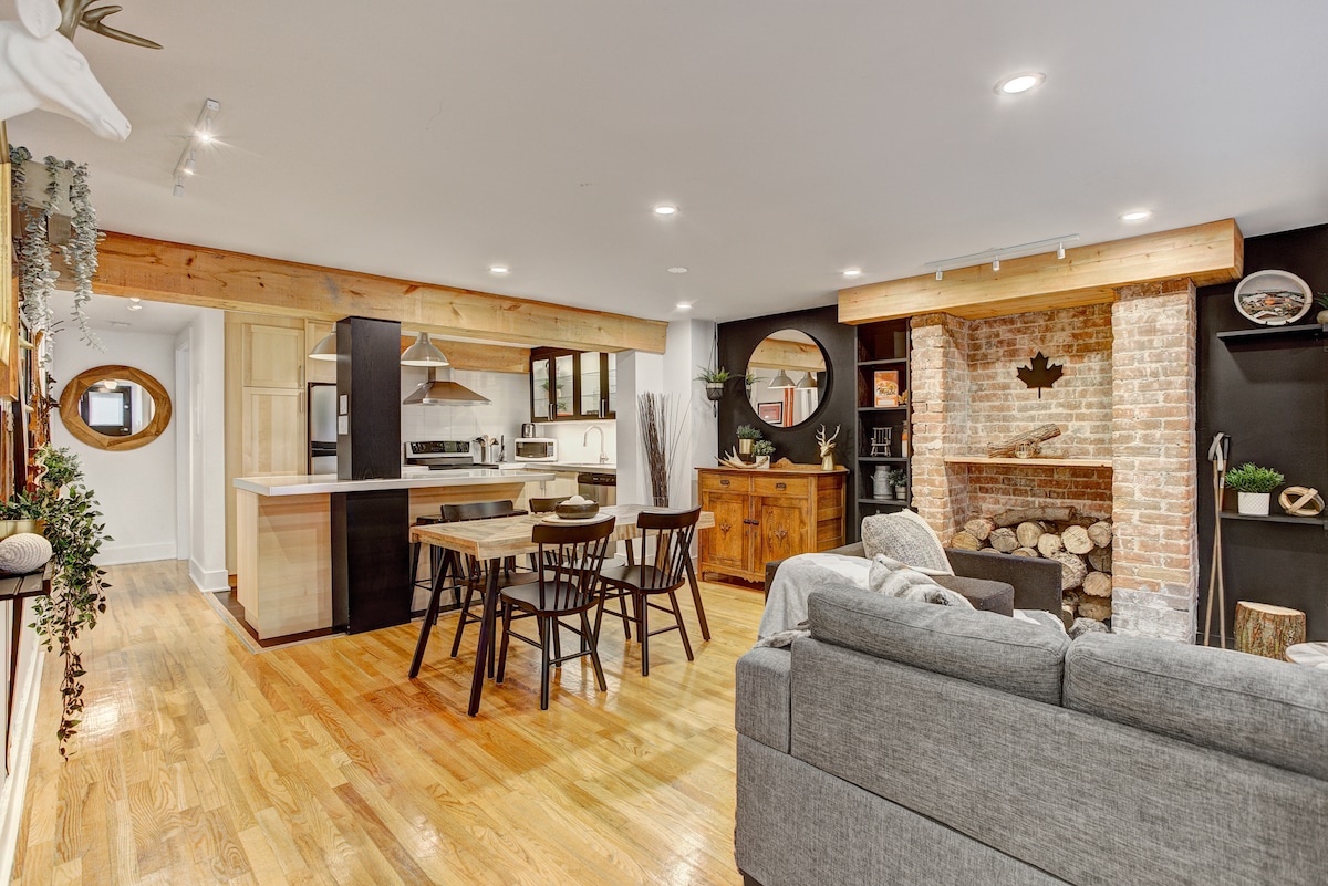An open living space is complemented by light wood accents and a cozy brick fireplace. A dining table with four chairs sits in the center, while a sofa is positioned nearby. The kitchen area features modern appliances and stylish cabinetry, enhancing the inviting atmosphere.