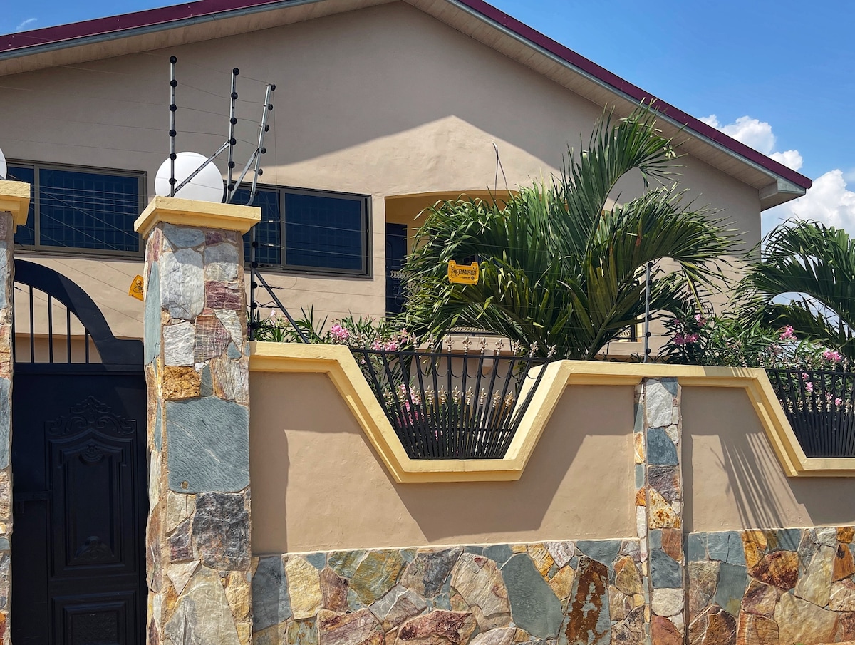 The exterior of the house features a stone wall with decorative elements and a gated entrance. Lush green plants are positioned along the fence, accompanied by light fixtures. Large windows allow natural light into the upper level, contributing to an inviting facade.