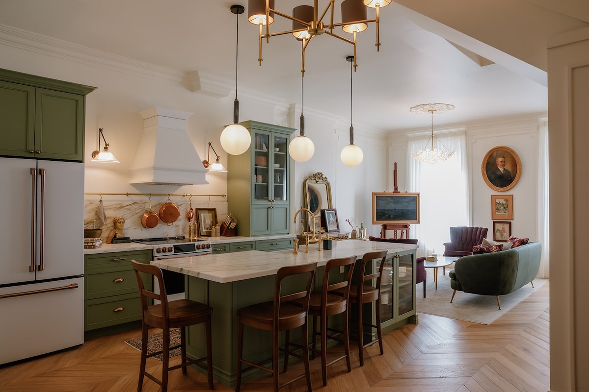 An open kitchen area features a large marble-topped island with six wooden bar stools. Green cabinetry complements the white marble backsplash and range hood. A vintage mirror and framed artwork add character to the space, while a chandelier with round lights hangs above, providing illumination.