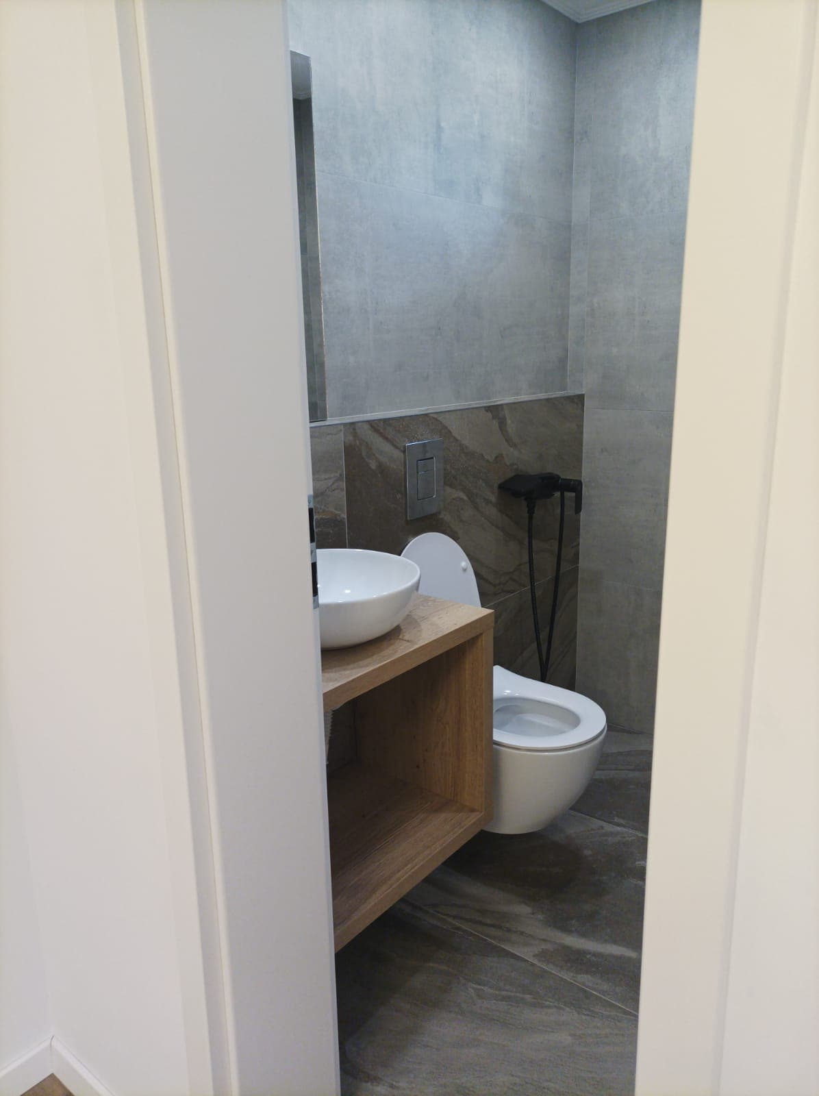 A modern bathroom features minimalist design elements. A round sink is set atop a natural wood vanity. The toilet is positioned adjacent, with gray stone tiles extending along the walls and floor, contributing to a clean and contemporary look.