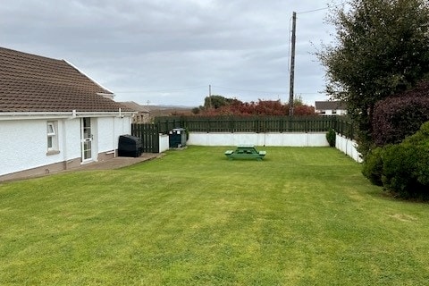 A spacious garden area is presented, featuring a well-maintained lawn that is bordered by a fence. A picnic table is positioned on the grass, accompanied by a charcoal grill. The cottage is visible in the background, showcasing white walls and a tiled roof.